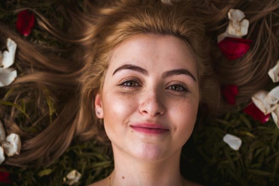 Junge Frau mit blonde Haaren, lächelnd auf grünem Hintergrund mit Blumenblättern.