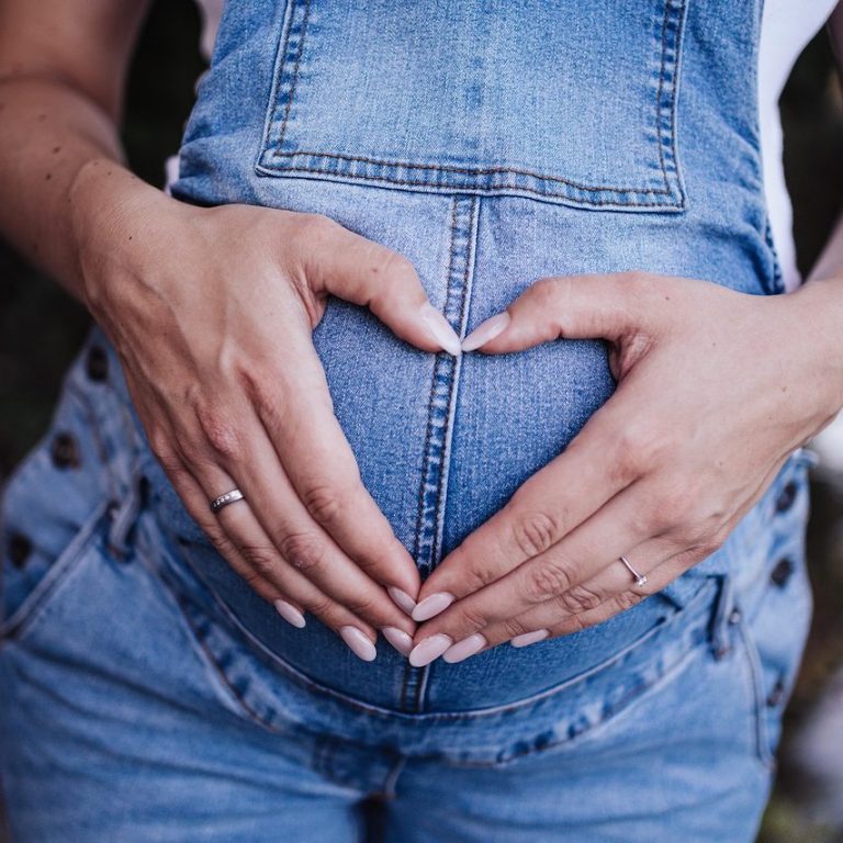 Schwangere Person formt mit Händen ein Herz auf ihrem Bauch.