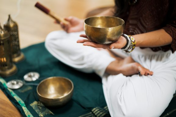 Person sitzt im Schneidersitz auf einer Yogamatte, mit einer Schale und einem Schlägel in den Händen.