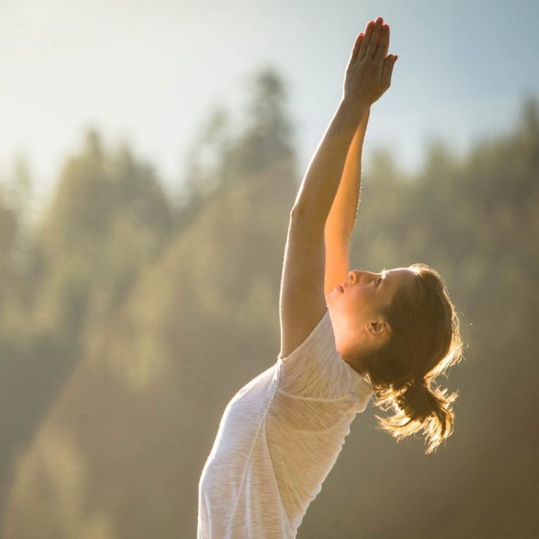 Frau in Yogapose mit erhobenen Händen vor einer bewaldeten Landschaft.