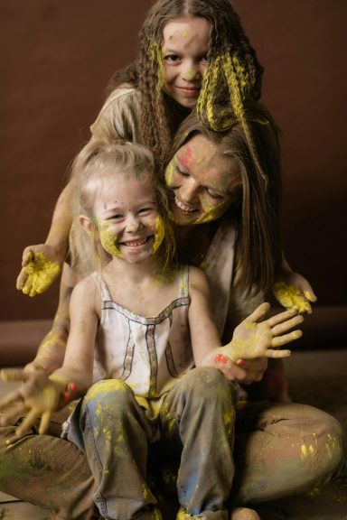 Eine Mama und ihre beiden Töchter posieren gemeinsam mit Farbe beschmiert und breitem Grinsen.