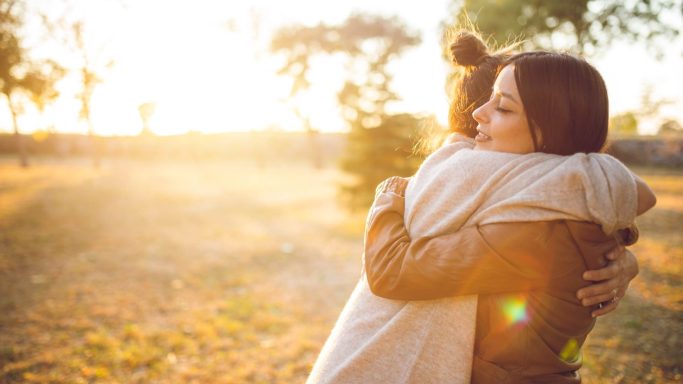 Zwei Frauen umarmen sich in einer sonnigen, natürlichen Umgebung.