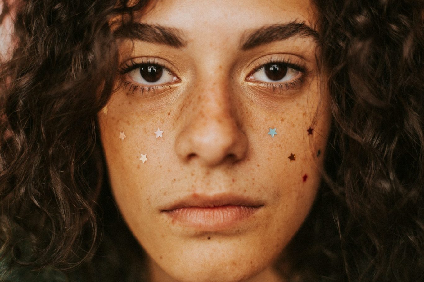Eine Person mit dunklen, lockigen Haaren und Sommersprossen, geschmückt mit bunten Glitzerpunkten.