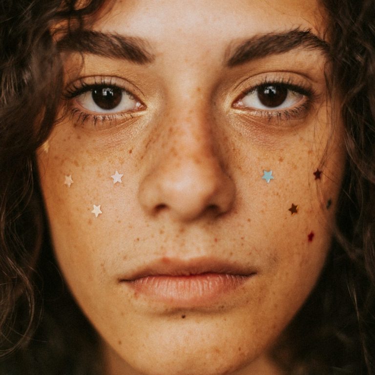 Porträt einer Frau mit lockigem Haar, Sommersprossen und bunten Glitzerpunkten im Gesicht.