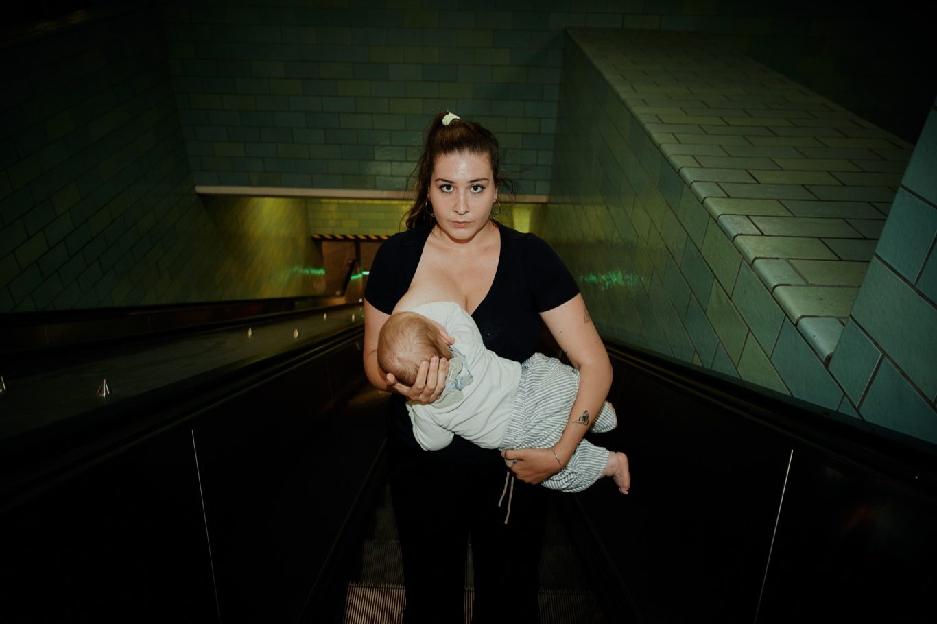 Foto: Lovis Trummer Photography Eine Frau stillt ihr Baby, während sie rückwärts eine Rolltreppe hinunterfährt.