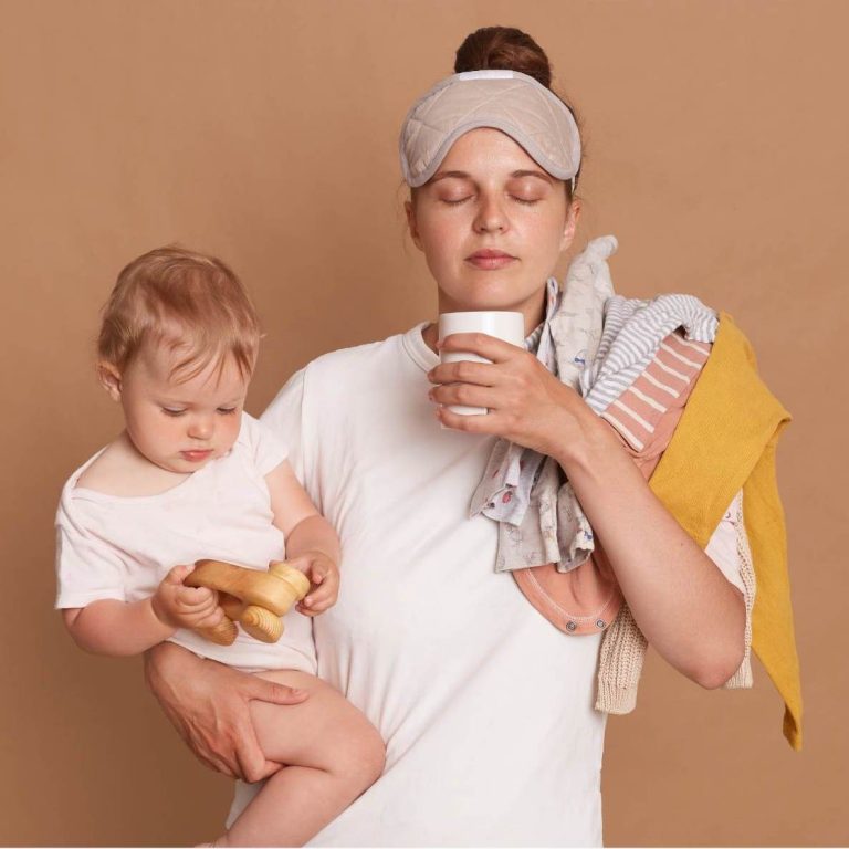 Frau mit Schlafmaske hält Baby und eine Tasse, unterstützt von Kleidung und einem Snack.