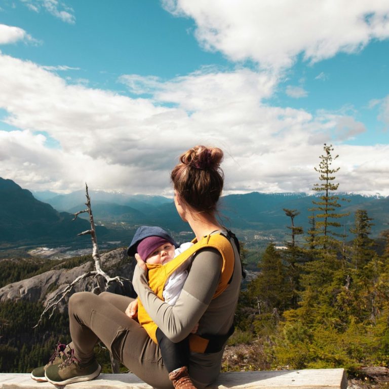 Frau mit Baby im Tragetuch blickt auf eine malerische Berglandschaft.