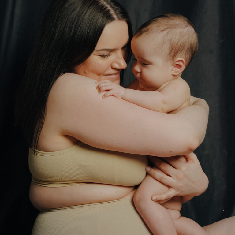 Eine Frau in Unterwäsche hält ein Baby in ihren Armen und lächelt liebevoll.