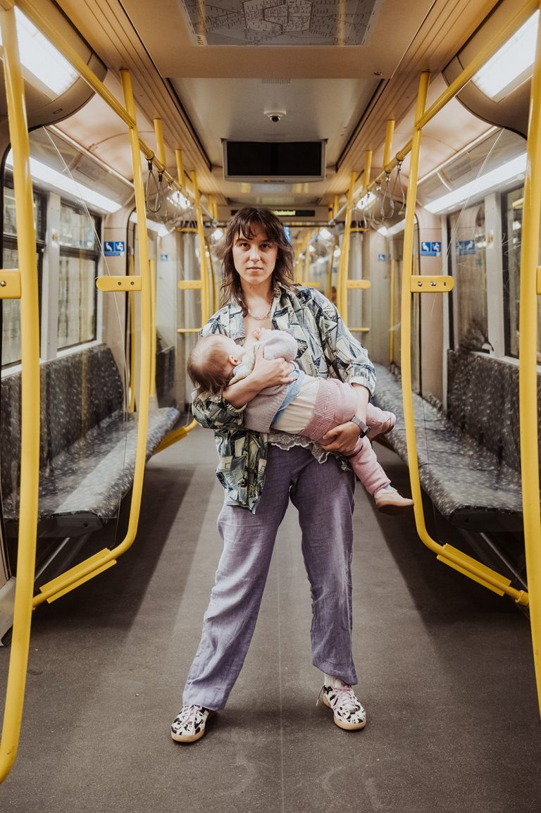 Foto: Magdalena Luise Mielke, Spiegelverdreht Frau stillt ihr Baby in der U-Bahn, umgeben von leeren Sitzplätzen.