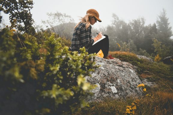 Frau in kariertem Hemd sitzt auf einem Stein und schreibt in ein Notizbuch in der Natur.