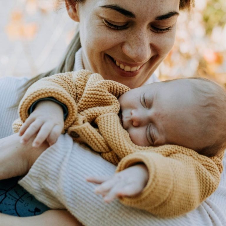 Eine lächelnde Frau hält ein schlafendes Baby in den Armen, umgeben von herbstlichen Pflanzen.