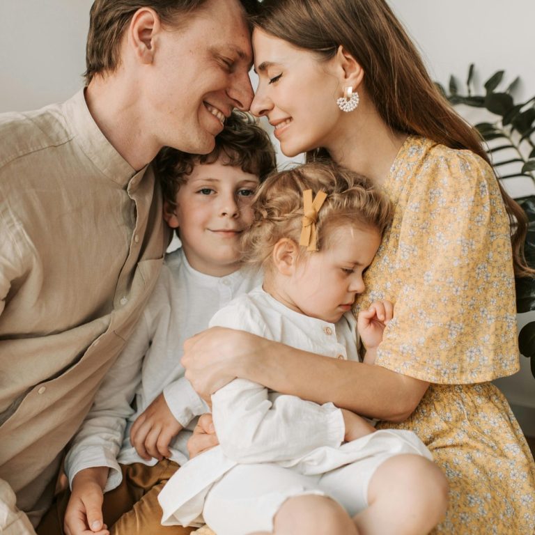 Familie posiert lächelnd zusammen, ein Junge und ein schlafendes Mädchen in den Armen der Eltern.