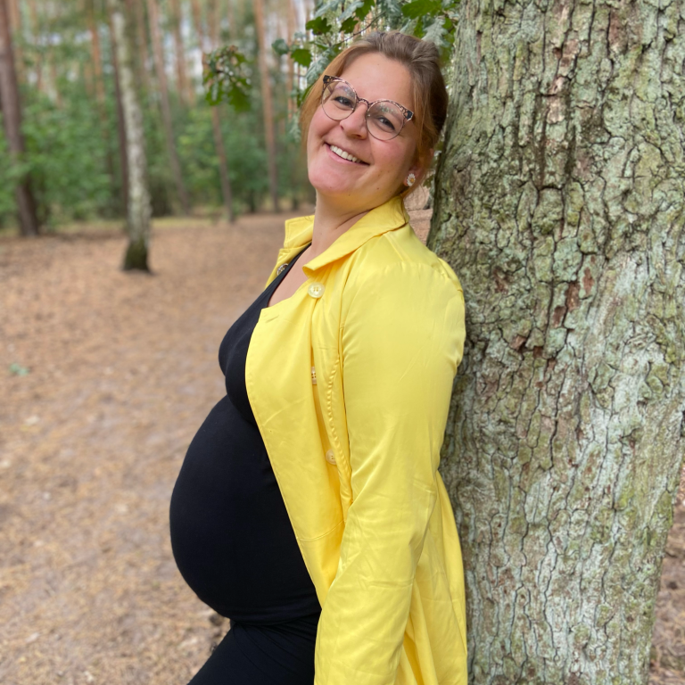 Schwangere Frau in gelbem Mantel lehnt lächelnd an einem Baum im Wald.