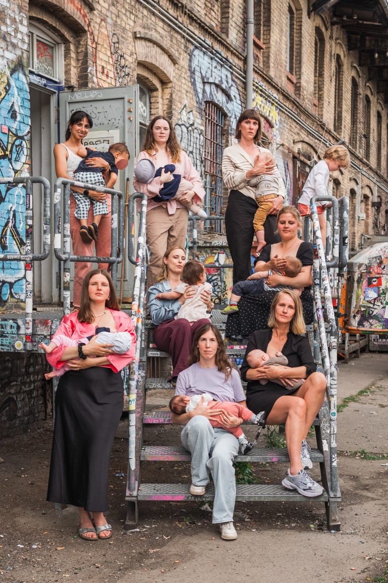 Foto: Una Oge, StudioUna Gruppierung junger Mütter beim Stillen ihrer Babys und Kinder auf einer Treppe vor einer graffitibedeckten Wand.