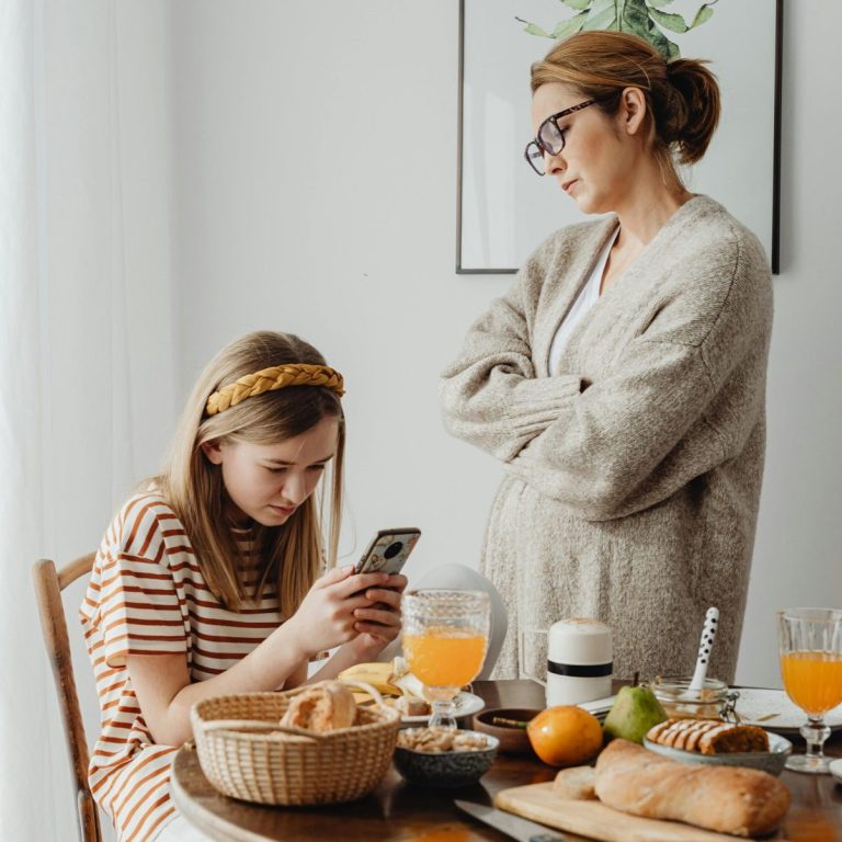 Ein Mädchen sitzt am Tisch und schaut auf ihr Handy, während ihre Mutter daneben steht.