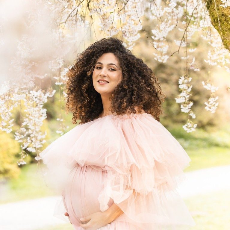 Schwangere Frau in einem rosa Kleid mit Volants vor blühenden Bäumen.