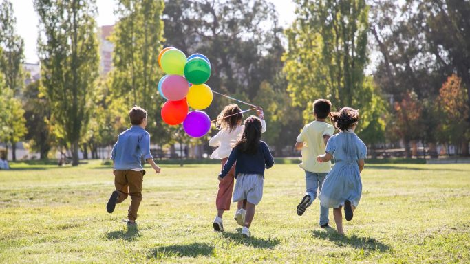 Fünf Kinder laufen fröhlich mit bunten Ballons über eine Wiese.