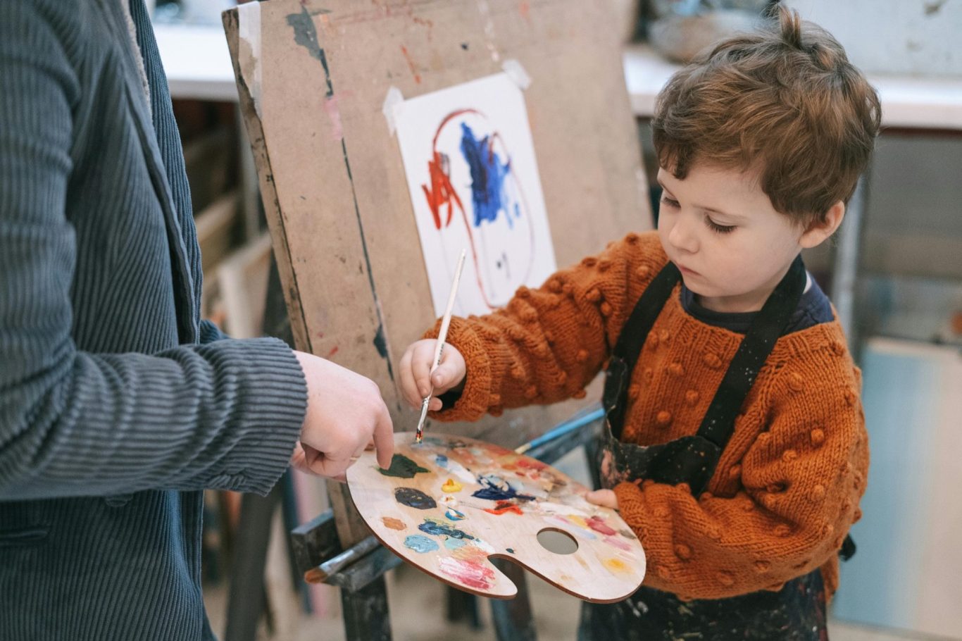 Ein Kind malt mit einer Farbpalette vor einer Leinwand in einem Atelier, die Mutter steht beratend daneben.