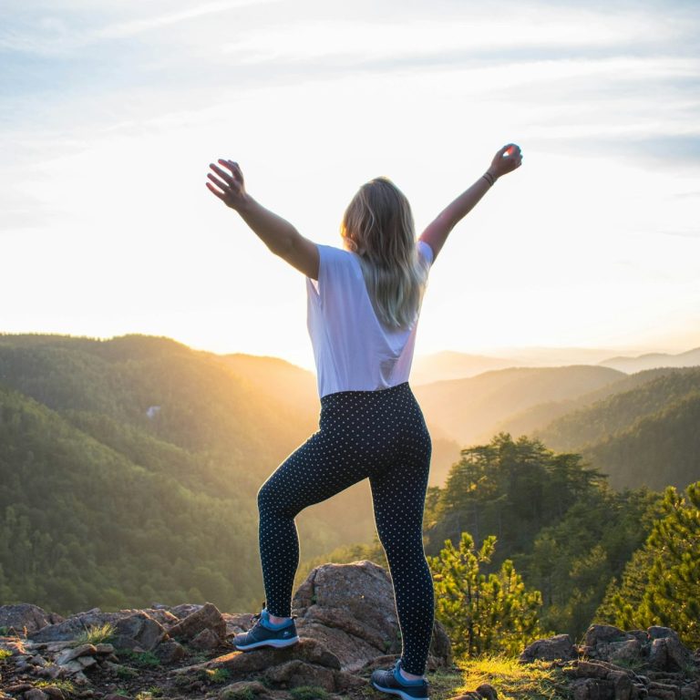 Frau steht auf einem Felsen, Arme ausgebreitet, mit Blick auf eine hügelige Landschaft bei Sonnenaufgang.