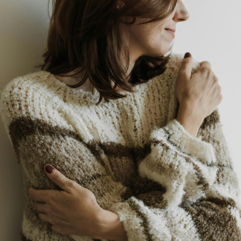 Frau mit braun-weißem Pullover, die nachdenklich an der Wand lehnt.