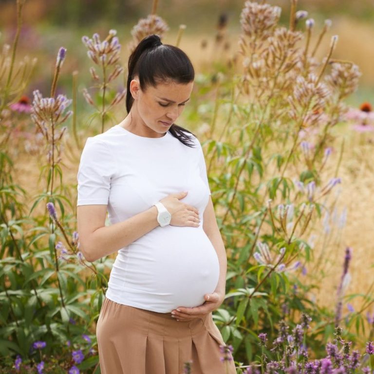 Schwangere Frau steht in einem Blumenfeld und hält ihren Bauch.