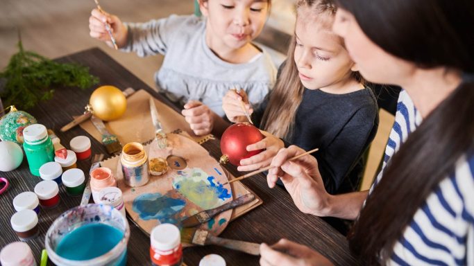Eine Mutter bastelt mit zwei Kindern bunte Weihnachtsbaumkugeln am Tisch.